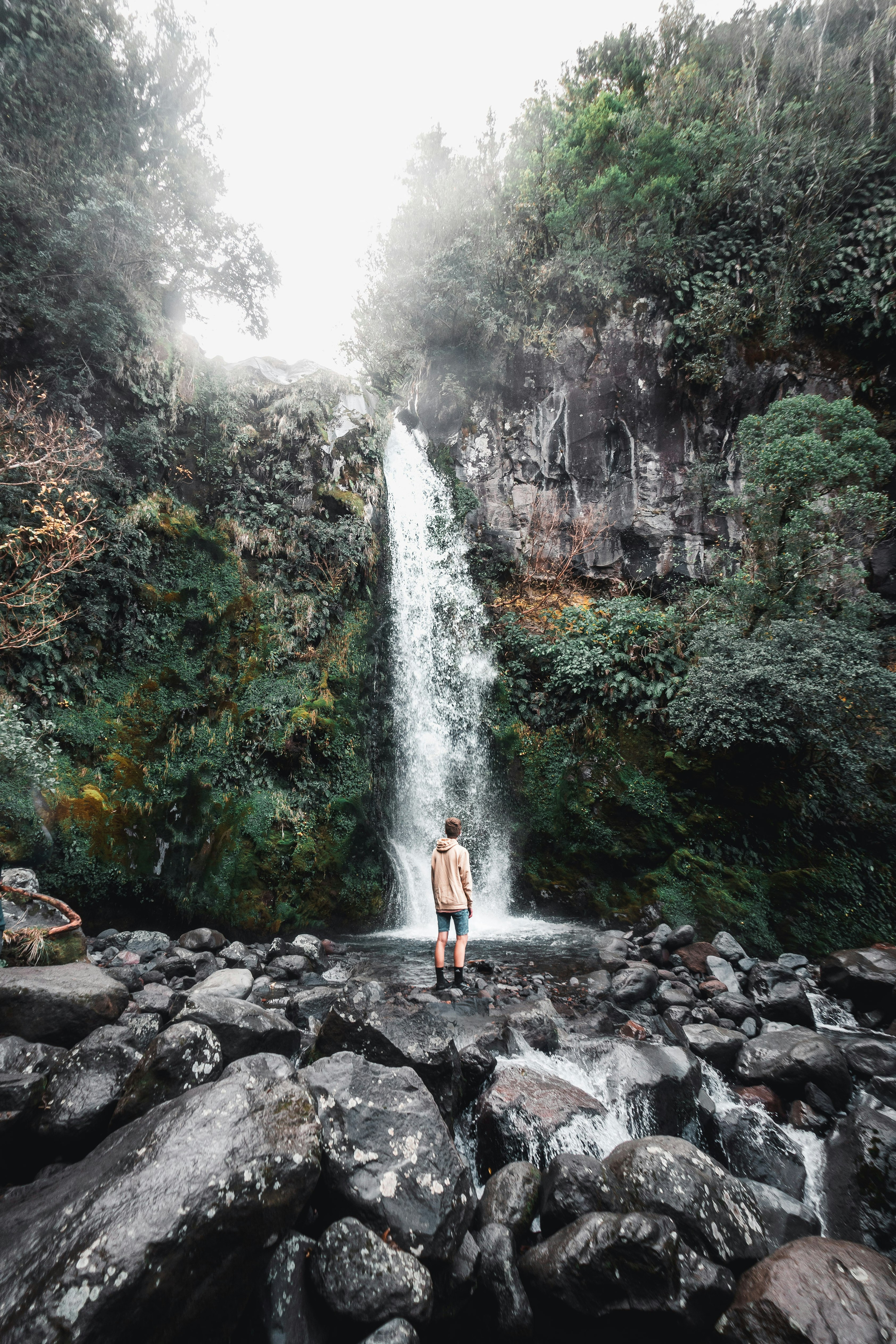 woman in white tank top standing on rocky shore in front of waterfalls during daytime